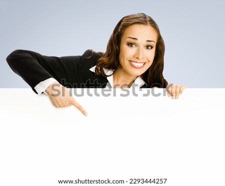 Happy excited smiling brunette business woman in blue clothing, standing behind, peeping from blank banner or mock up signboard, showing pointing forefinger copy space area for text, grey background.