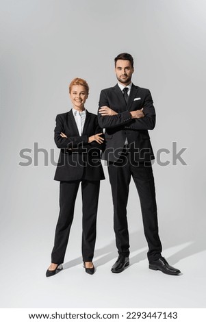 full length on successful business partners in black suits crossing arms and looking at camera on grey background