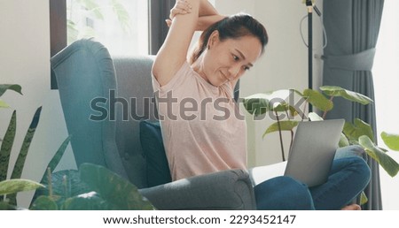 Young Asian woman wear casual sitting on wing chair use laptop computer focus on screen typing report feel tired stretching arm raise hand with plant in living room greenhouse. Work from home concept.