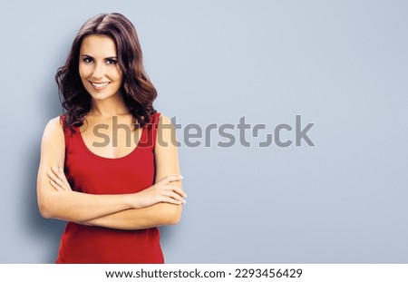 Portrait photo - smiling woman in casual red clothing, in crossed arms pose. Grey background. Happy beautiful girl at studio. Brunette model with long hair posing at studio picture.