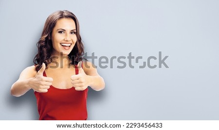 Portrait photo - happy smiling woman in casual clothing, showing thumbs up gesture, over grey color wall background. Girl in red dress. Brunette excited model at studio picture.
