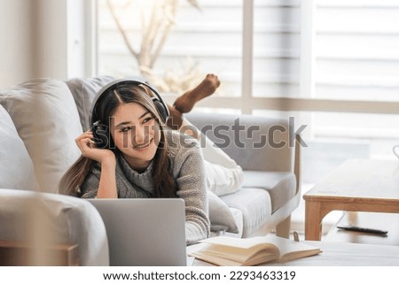 Photo of modern asian woman wearing headphones use laptop while lying on sofa in bright home