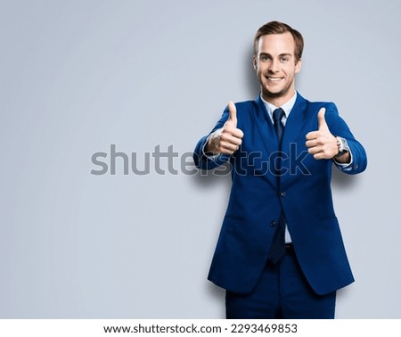 Cheerful smiling businessman in blue confident suit showing two thumbs up like hand gesture, over grey color background with copy space for ad, slogan or text. Successful business man.
