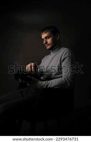 Portrait of a young handsome man in a gray jacket with a camera in his hand. The photographer on a black background sits on a chair. classical portrait