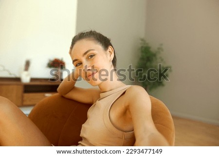 Cute girl taking selfie for dating application profile picture, looking at camera with happy friendly smile, dressed in beige summer tank top, sitting in brown armchair in minimalistic living room Royalty-Free Stock Photo #2293477149