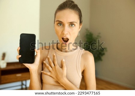 Shocked surprised female of 20s in tank top showing black blank copy space screen of smartphone putting hand to chest with mouth opened with surprise, saying omg, excited with special offer or sales