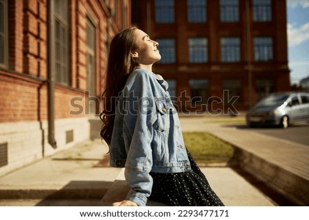Side view outdoor image of gorgeous student girl with closed eyes in denim outfit leaning on edging at city streets taking sun bathing with face turned to sunlight, enjoying warm day and freedom Royalty-Free Stock Photo #2293477171