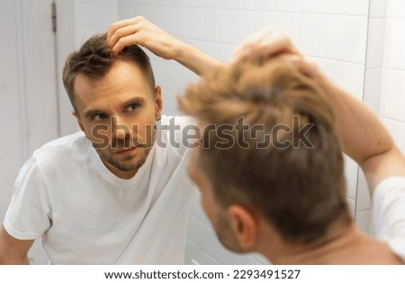Middle aged caucasian white man with a short beard looks at his hair in the mirror in the bathroom and worried about balding. The concept of the problem of male hair loss, early baldness and alopecia. Royalty-Free Stock Photo #2293491527