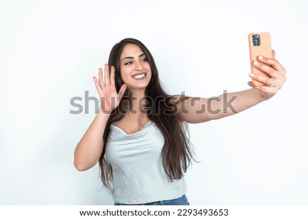young brunette woman wearing white tank top over white studio background holds modern mobile phone and makes video call waves palm in hello gesture. People modern technology concept
