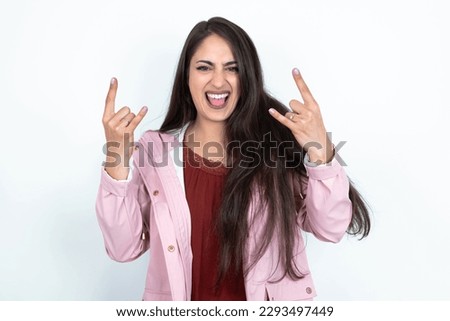 young brunette woman wearing pink raincoat over white studio background makes rock n roll sign looks self confident and cheerful enjoys cool music at party. Body language concept. Royalty-Free Stock Photo #2293497449
