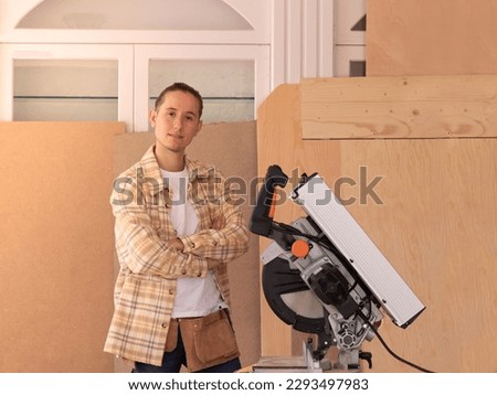 Young male carpenter with arms crossed standing in home carpentry shop against wood planks near modern mitre saw