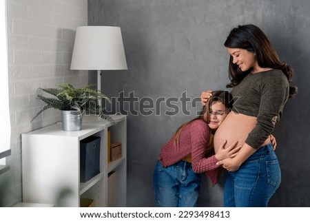 Happy young girl leaning head on mother's pregnant belly and listening baby's heartbeat. Happy little girl feeling baby at mom's tummy and awaiting the birth of her little brother.