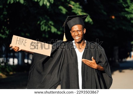 Portrait of smiling black guy standing and pointing at cardboard poster on street in sunny day looking for job, employment issue. University or college graduating student in graduate gown and cap. Royalty-Free Stock Photo #2293503867