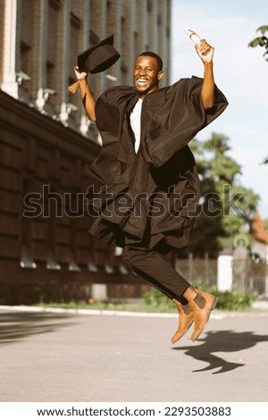 Happy black guy graduate from university with pleasure jumping up in black mantle and hat. Photo of student outdoors on sunny day. Higher education diploma, future career, graduation, celebration.