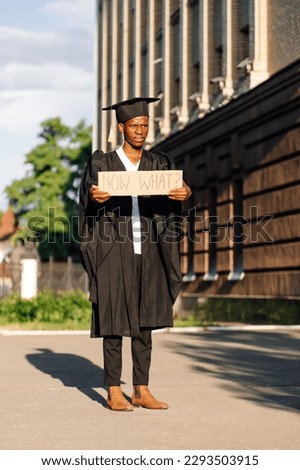Dissatisfied afro american guy standing with cardboard poster on street in sunny day looking for job. University or college graduating student in graduate gown and cap. Employment issue concept.  Royalty-Free Stock Photo #2293503915