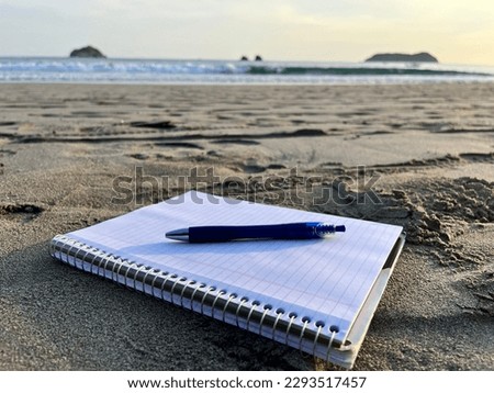 A coiled scribbler notebook with a pen on it on a beach with the sky in the background as a concept image for relaxation and journaling Royalty-Free Stock Photo #2293517457