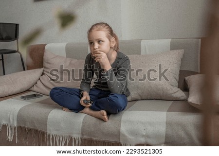 A cute little girl is sitting on the couch at home, using the remote control to switch channels on the TV. Watch informative and interesting children's programs or videos for children and teenagers