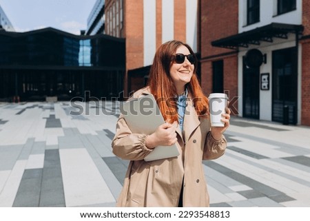 Caucasian redhead businesswoman in fashion office clothes posing outside with a laptop and eco cup of tea. Mockup, Woman using of the notebook computer and holding with bio coffee cup