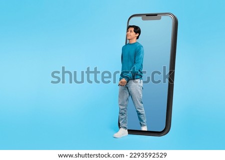Asian Teen Guy Walking Out Of Large Phone Screen Looking Aside At Copy Space Posing On Blue Background In Studio. Student Advertising Mobile Offer Or App. Full Length, Collage