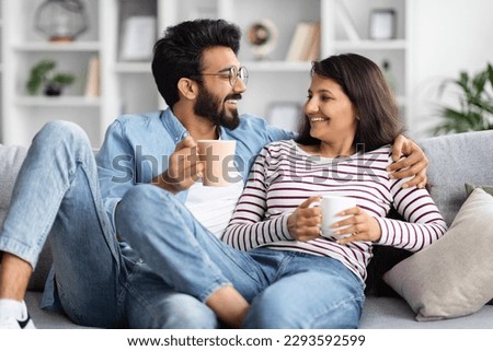 Relaxed happy hindu young lovers with tea mugs sitting on couch in living room, drinking coffee and having conversation, eastern man and woman enjoying time together at home Royalty-Free Stock Photo #2293592599