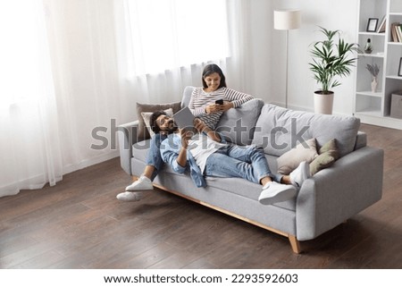 High angle view of relaxed cheerful stylish millennial eastern lovers man and woman chilling on couch in cozy living room, using gadgets at home, scrolling on social media, reading online, copy space Royalty-Free Stock Photo #2293592603