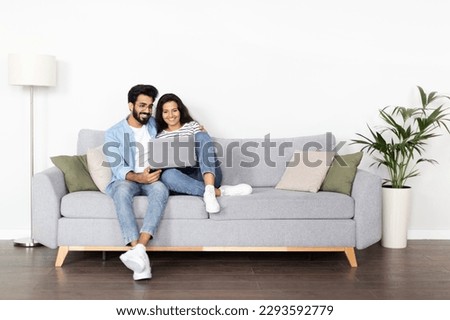Loving happy beautiful eastern couple in stylish casual outfit sitting on couch at home, embracing, using laptop, young indian man and woman watching movie online, copy space, full length Royalty-Free Stock Photo #2293592779