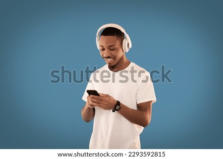 Cool happy smiling young african american guy in casual using brand new wireless headphones and smarphone on blue studio background, watching video content online, copy space. Modern technologies Royalty-Free Stock Photo #2293592815
