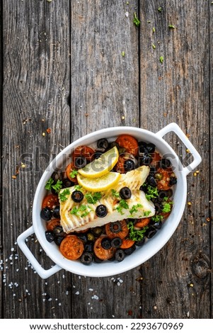 Cod in Italian style on tomatoes and black olives with baked potatoes in baking dish on wooden table 