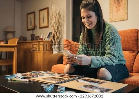 Middle-aged brunette woman sitting on her living room sofa holding and looking at her smartphone while smiling and making her handmade kraft travel album with washi tape. Royalty-Free Stock Photo #2293672031