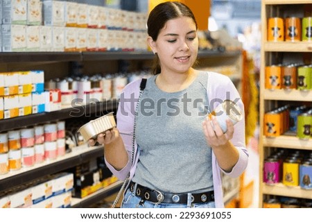 Portrait of interested young woman carefully reading labels on tin cans with choosing right hypoallergenic food for her dog in pet store Royalty-Free Stock Photo #2293691055