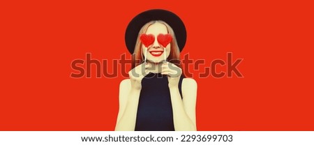 Portrait of beautiful young woman covering her eyes with red heart shaped lollipop wearing black round hat on background