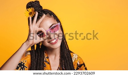 Summer girl in sunglasses and sunflower in her hair, shows zero, okay sign and smile, holiday and vacation concept, stands in bright dress isolated on yellow background.