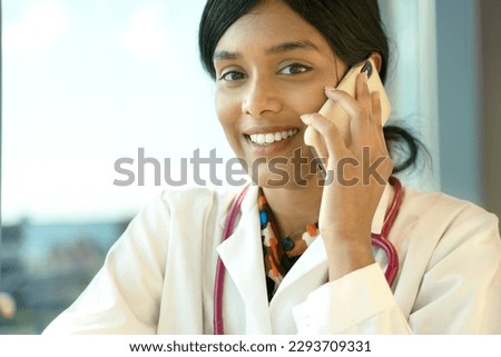 Portrait of smiling Indian female student wearing white coat and stethoscope, looking at camera. Beautiful female doctor talking on mobile phone in hospital. Medicine, health care
