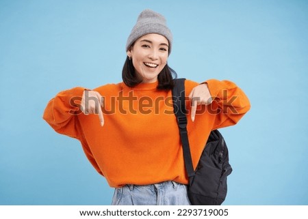 Asian woman in warm hat, wears street outfit and backpack, points fingers down, shows advertisement below, standing over blue background.