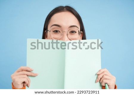 Portrait of cute japanese woman in glasses, peeking from notebook, covers face with her notes, smiles with eyes, blue background.
