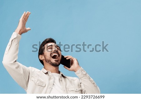 Portrait of a man brunette animation and joy talking on the phone hand and fist up from victory joy, on a blue background in a white T-shirt and jeans, copy space