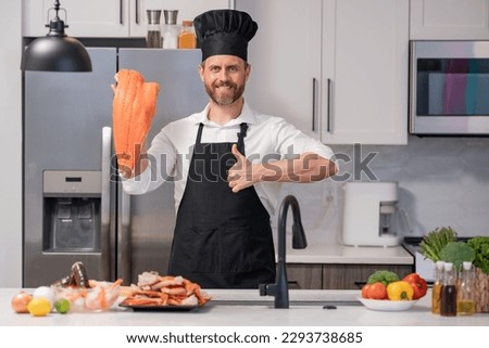 Portrait of cook man preparing fresh salmon at kitchen. Handsome man in cook apron and chef hat preparing raw fish salmon. Chef cooking seafood in kitchen. Millennial man hold raw fish salmon.