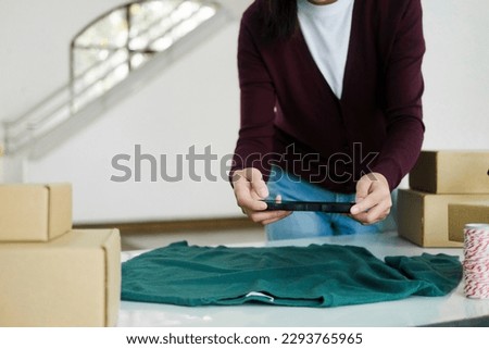 Close up of young female business owner in the standing at table with cardboard boxes in workplace taking picture of clothes, product using smartphone to post and sell in online website. 