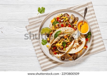 Grilled Steak Tacos with olives, tomatoes, red onion, avocado, cilantro, corn, shredded cheddar cheese and thousand island sauce on platter on white wood tabe, flat lay, free space