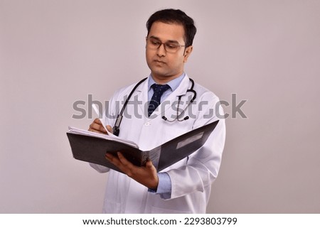 Indian male doctor pediatric, physical, therapist wearing white medical gown with stethoscope on shoulders stands writing in documents file isolated on grey. Healthcare and medicine
