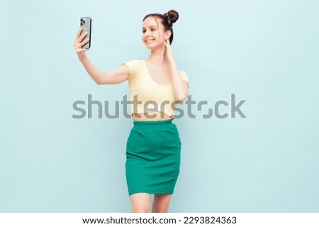 Young beautiful smiling female in trendy summer yellow t-shirt and green skirt. Carefree woman with two horns hairstyle posing near blue wall in studio. Positive model having fun. Taking selfie photos