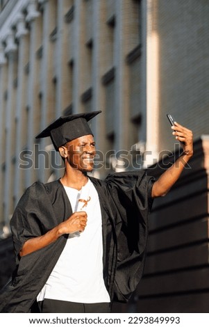 Smiling african american graduate from university taking photo with front camera of smartphone with higher education diploma. Student in black mantle and hat. Future career, graduation, celebration.