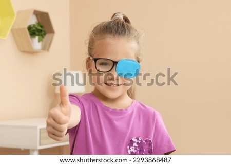 Girl with eye patch on glasses showing thumb up in room. Strabismus treatment Royalty-Free Stock Photo #2293868487