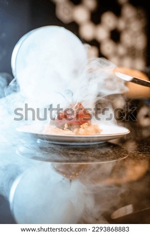 Presentation of an appetizer with a glass dome and interior smoke, inside a plate of an appetizer for the enjoyment of the diners. Black background and reflective table. Product photo