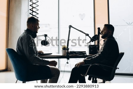 Two college podcasters laughing and having a good time in a studio. Two happy young men co-hosting a live audio broadcast. Two male content creators recording an internet podcast.