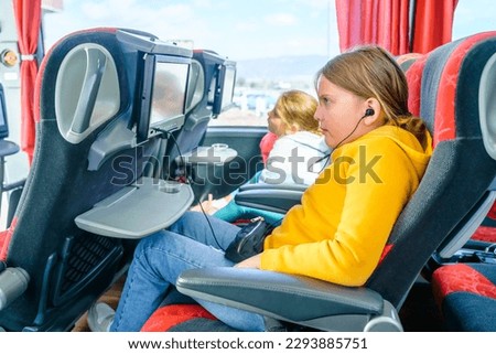 Bus road traveling. Girl kid rides on large comfortable sightseeing excursion bus. Watching movie cartoon on screen in chair. Entertainment media system. Sittting, listening to music in headphones.