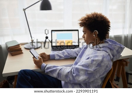 Youthful African American boy in earphones looking at smartphone screen while playing game or choosing music from playlist against laptop Royalty-Free Stock Photo #2293901191