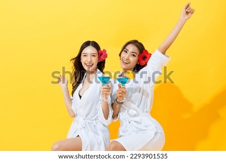 Two beautiful Asian woman friends wearing spa bathrobe holding summer cocktail drinks in yellow color isolated background studio shot