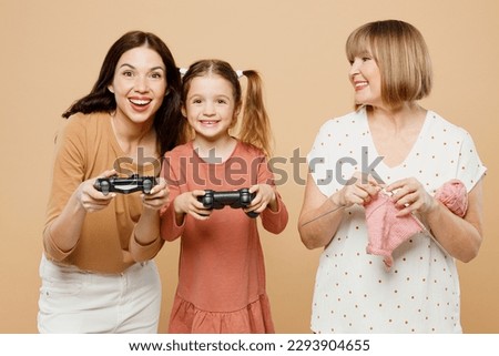 Happy women wears casual clothes with child kid girl 6-7 years old. Granny mother daughter knit hold play pc game with joystick console isolated on plain beige background. Family parent day concept