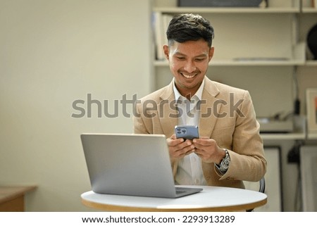 Smiling businessman in stylish suit working with laptop and using smartphone in modern office space